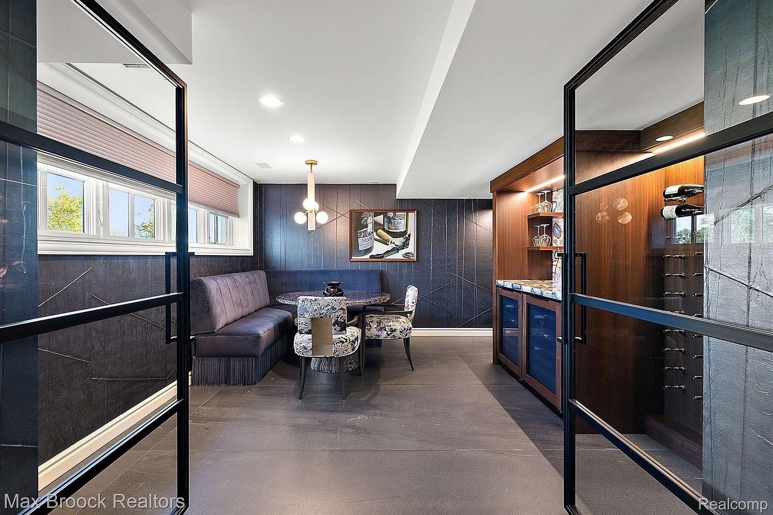 This interior shot showcases a stylish dining area with a built-in booth seating arrangement and two patterned chairs around a round table. The room features dark, textured walls, a modern chandelier, and a built-in bar area with wine storage, creating a sophisticated and inviting space. The view is from the doorway, giving a sense of entry into the room.
