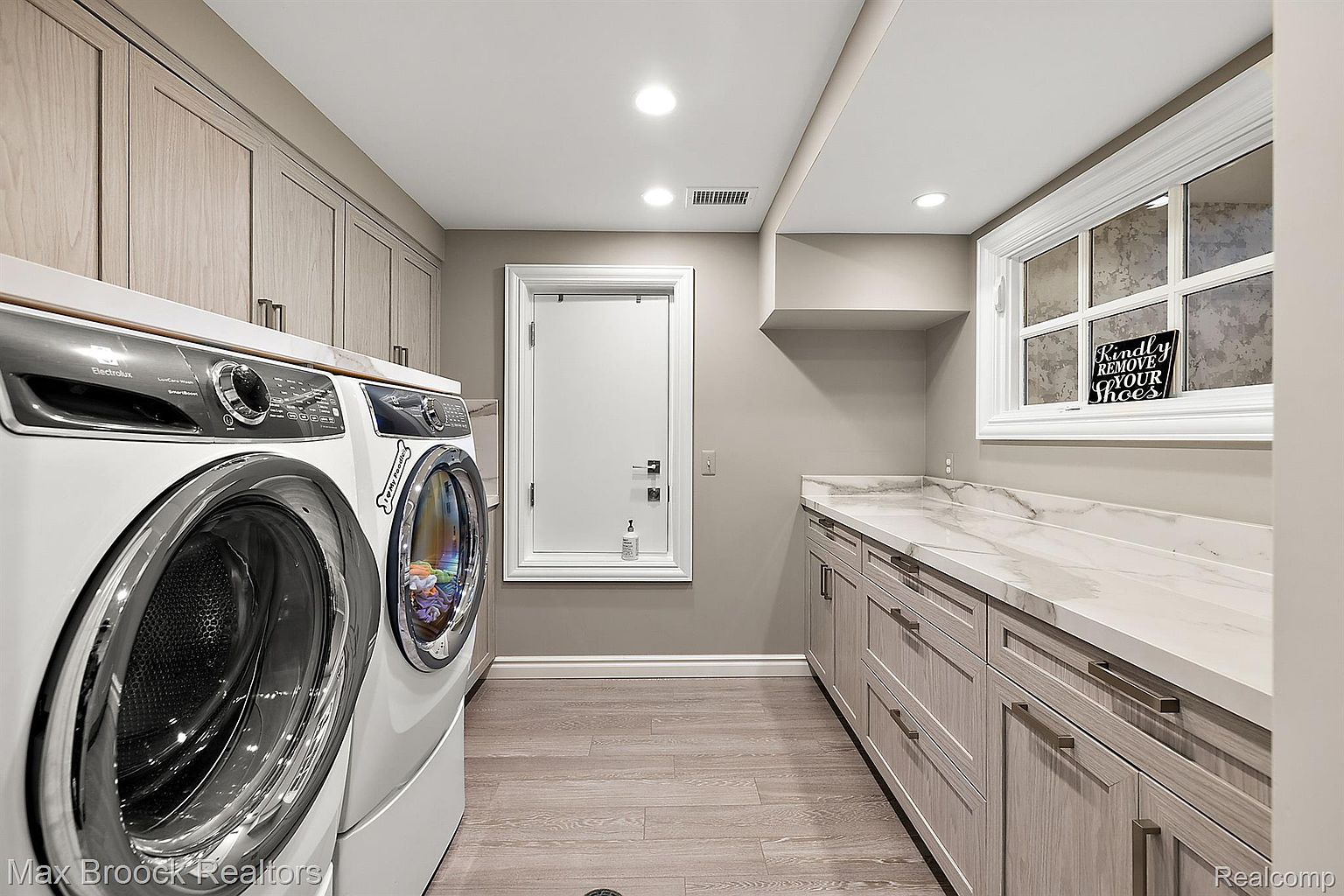 This is a well-organized laundry room featuring a front-loading washer and dryer set, light wood cabinetry with a countertop, and a window providing natural light. The room has a clean and modern aesthetic with light-colored walls and wood-look flooring, creating a functional and appealing space.