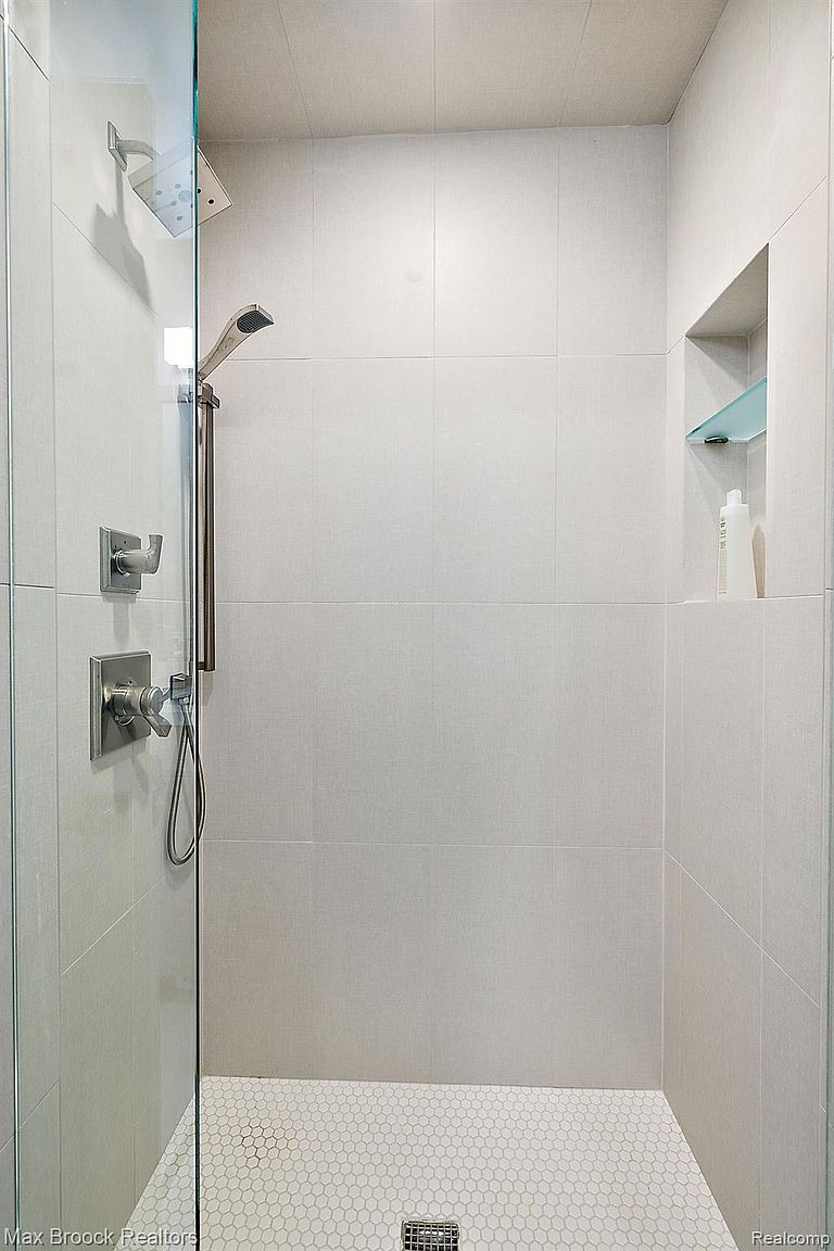 This is a well-lit, modern shower featuring white tile walls and a white hexagonal tile floor. The shower has a glass door, a rainfall shower head, and a handheld shower wand. A built-in niche with a glass shelf holds toiletries, adding a touch of convenience and style.