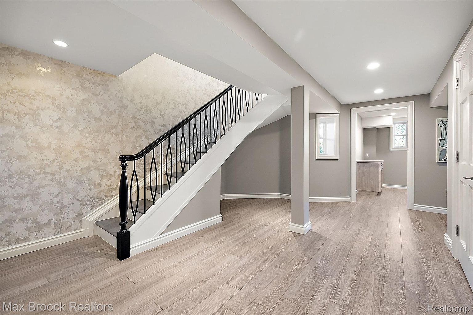 This is a well-lit basement interior featuring light wood-look flooring and neutral gray walls. A staircase with a black wrought iron railing leads to the upper level. The space includes a support column and a doorway leading to another room, suggesting a finished basement suitable for various uses.