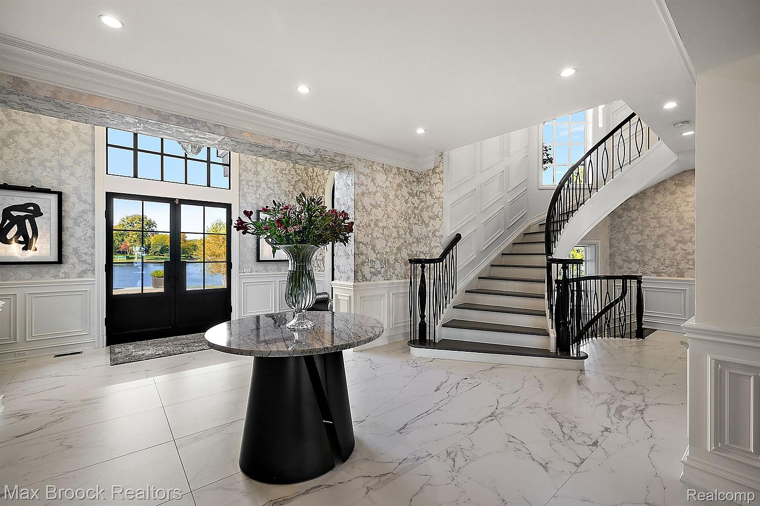 This grand foyer features a stunning curved staircase with black wrought iron railings and dark wood treads, complemented by elegant white marble flooring. A round table with a floral arrangement sits in the center, adding a touch of sophistication, while the large windows and doors provide ample natural light and a view of the outdoors. The walls are adorned with decorative wallpaper and wainscoting, enhancing the luxurious feel of the space.