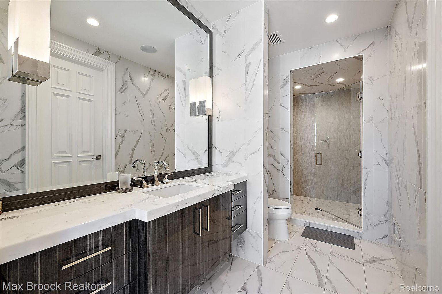 This is a luxurious primary bathroom featuring marble-like tile on the walls and floors, creating a seamless and elegant look. The vanity has a dark wood finish with a white countertop and modern fixtures. A glass-enclosed shower is visible in the background, and the overall impression is clean, bright, and upscale.