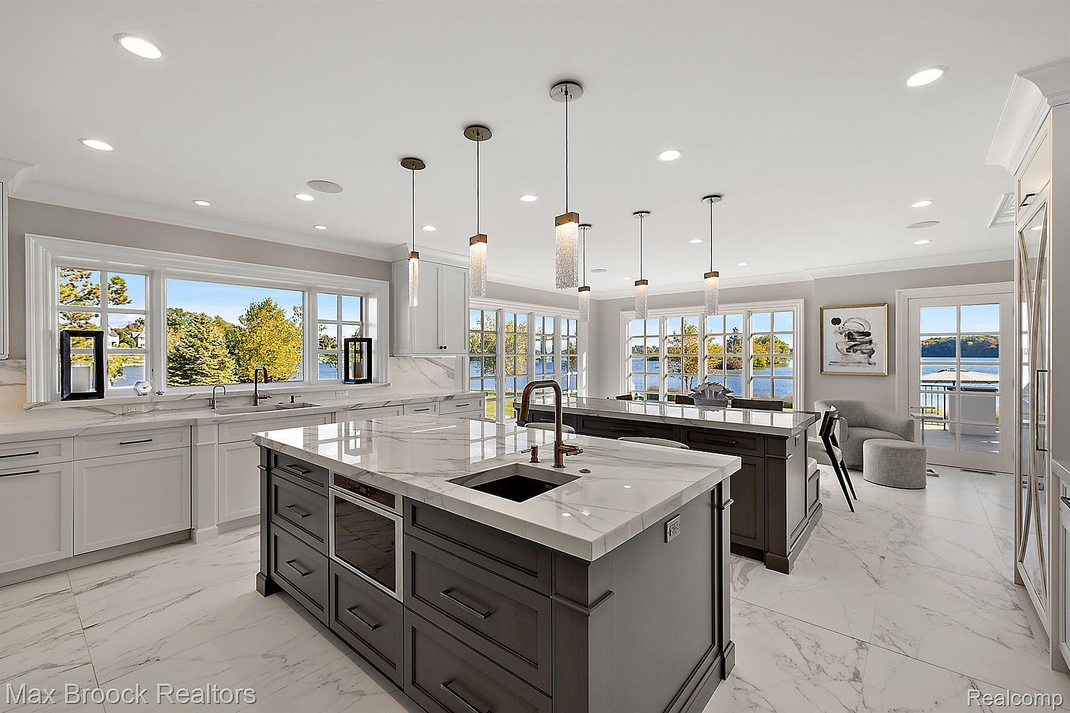 This is a bright and spacious kitchen featuring two large islands with marble countertops and dark wood cabinetry. The kitchen boasts white cabinets along the perimeter, stainless steel appliances, and multiple windows offering scenic water views. Modern pendant lighting hangs above the islands, enhancing the luxurious feel of the space.