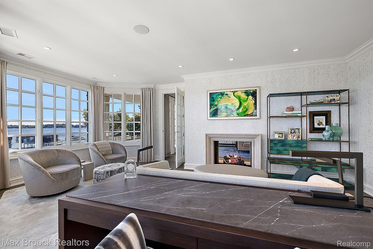 This interior shot showcases a sophisticated office space with a large desk featuring a dark marble top, complemented by comfortable seating and a view of the water through expansive windows. A fireplace with a modern art piece above it and a stylish shelving unit add to the room's elegance and functionality, creating an inviting and productive atmosphere. The room is well-lit with natural light and recessed lighting.
