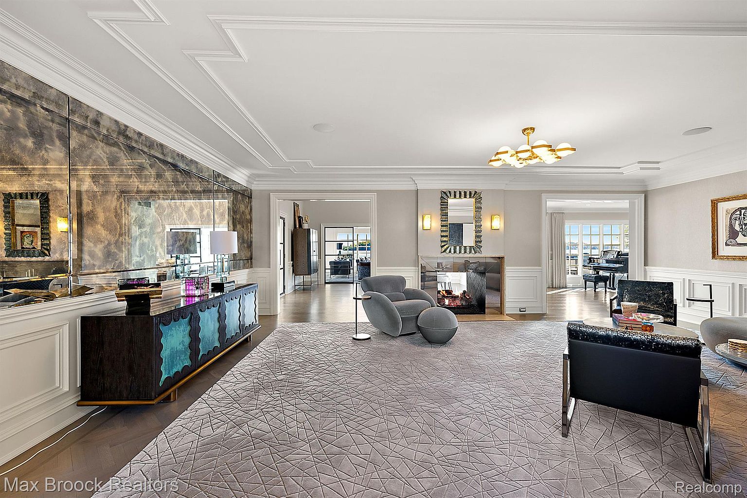 This is a spacious living room featuring a large patterned rug, a modern fireplace, and elegant furnishings. The room is well-lit with a combination of natural light and stylish ceiling fixtures. A mirrored wall adds depth and reflects the light, enhancing the room's luxurious feel.