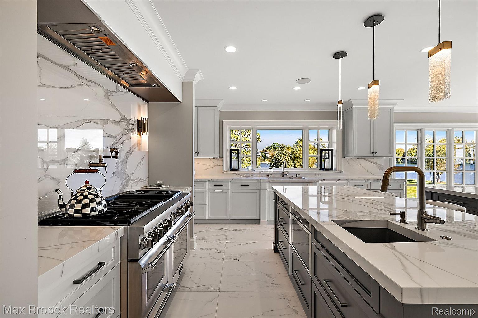 This is a bright and modern kitchen featuring white marble countertops and backsplash, stainless steel appliances, and a large island with a sink. The kitchen is well-lit with recessed lighting and pendant lights, and large windows offer a view of the outdoors. The overall impression is one of luxury and sophistication.
