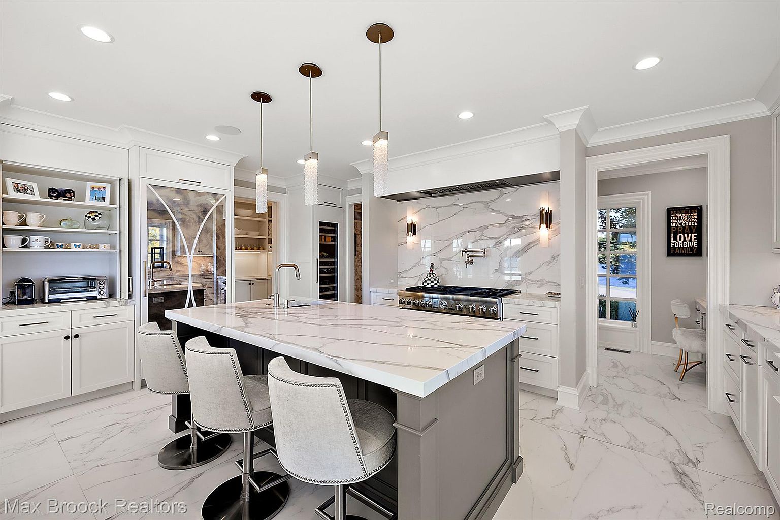 This is a bright and luxurious kitchen featuring a large island with a marble countertop and seating. The kitchen is equipped with high-end appliances, custom cabinetry, and elegant pendant lighting. The open layout and marble flooring contribute to a sophisticated and modern aesthetic.