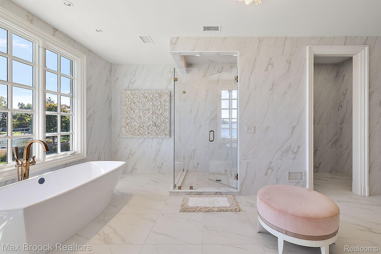 This is a luxurious primary bathroom featuring marble tile throughout, a freestanding soaking tub with a gold faucet, and a glass-enclosed shower. A pink ottoman adds a touch of elegance, and a large window provides natural light and a view. The overall impression is one of spa-like tranquility and high-end design.