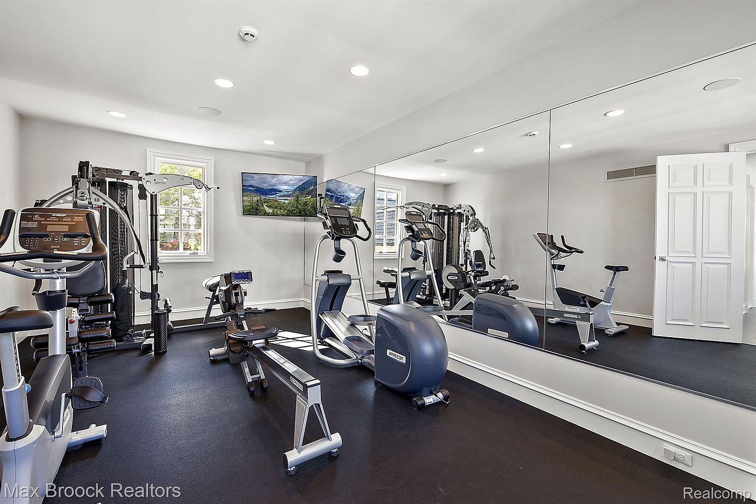 This is a well-equipped home gym featuring various exercise machines, including a multi-station weight machine, elliptical, rowing machine, and stationary bike. The room has a large mirror, a window providing natural light, and two wall-mounted televisions. The floor is covered in a dark rubber material, and the walls are painted a neutral color, creating a clean and functional workout space.