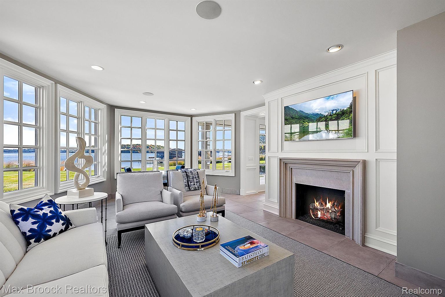 This is an interior shot of a luxurious living room featuring a fireplace with a television mounted above it. The room is well-lit with natural light from large windows offering a water view, and it includes comfortable seating arrangements with a sofa and armchairs. The overall impression is one of elegance and comfort, perfect for relaxation and entertaining.