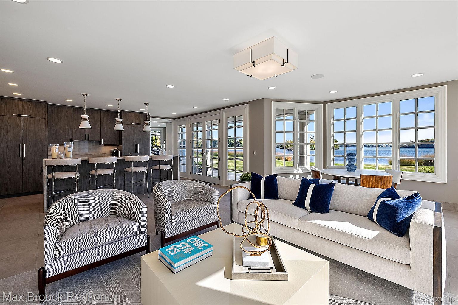 This is an interior shot of a modern living room that seamlessly blends into a kitchen area. The living room features a comfortable sofa with blue pillows, two armchairs, and a stylish coffee table with decorative objects. Large windows offer a beautiful view of the water, enhancing the room's appeal and creating a bright, airy atmosphere.