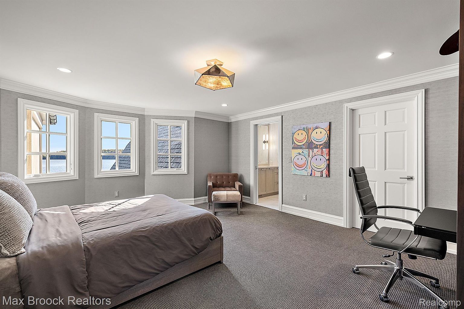 This is a primary bedroom featuring a neutral color palette with gray walls and carpet. The room has three windows offering natural light and a view of the water. A bed with gray bedding is the focal point, and there is a seating area with a chair and ottoman. A desk and chair are positioned near a door, and artwork adorns the wall, adding a touch of personality.