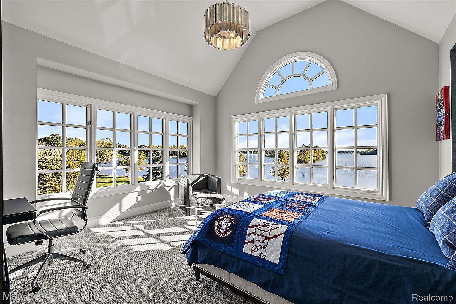 This is a bright and airy primary bedroom featuring large windows with views of the water. The room is painted in a neutral gray tone, complemented by a modern chandelier and a blue bedspread. A desk and chair are positioned near the windows, creating a functional workspace.