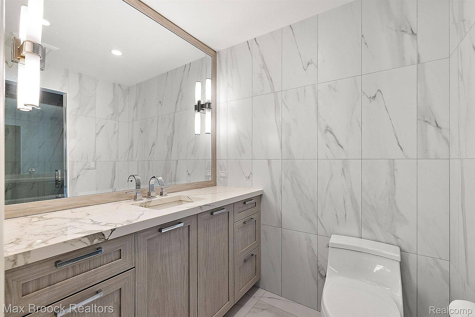 This is a well-lit primary bathroom featuring a double vanity with a light wood finish and a white marble countertop. The walls are covered in large-format marble tiles, creating a luxurious and clean aesthetic. A large mirror reflects the vanity and the opposite wall, enhancing the sense of space, and a modern toilet is visible to the right.
