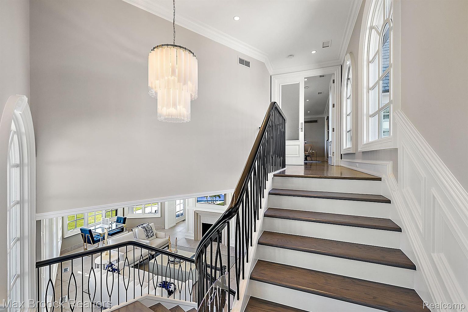 This interior shot showcases a grand hallway with elegant stairs. The stairs feature dark wood treads and white risers, complemented by a black wrought iron railing. A modern chandelier hangs above, and arched windows provide natural light, creating a bright and inviting space.
