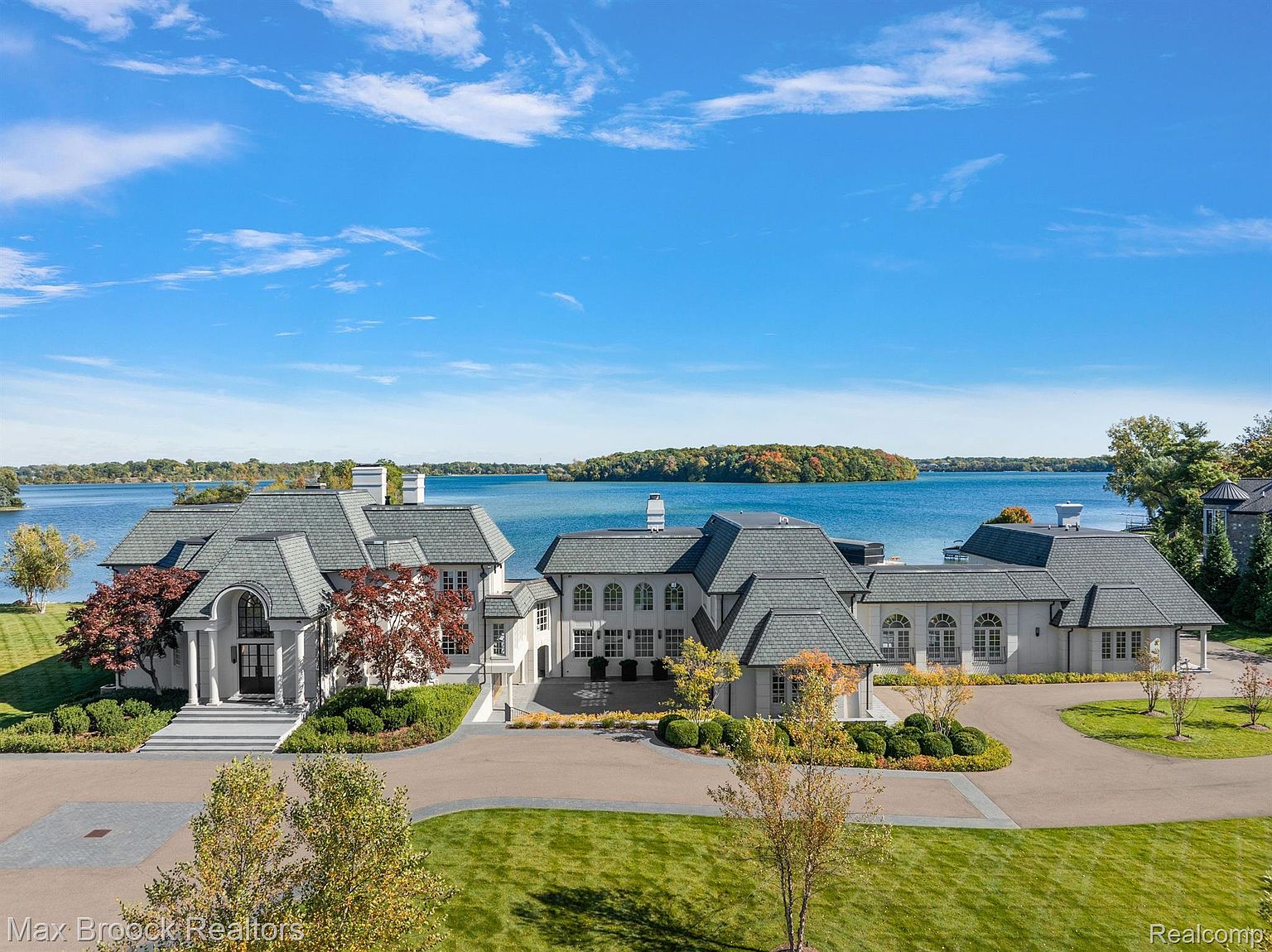 This aerial view showcases a grand estate with multiple interconnected structures, featuring a cohesive architectural style with gray roofs and light-colored exteriors. The property is meticulously landscaped with lush green lawns, mature trees, and manicured shrubbery. The estate is situated on a waterfront property with a clear blue lake and a small island in the background, creating a serene and luxurious setting.