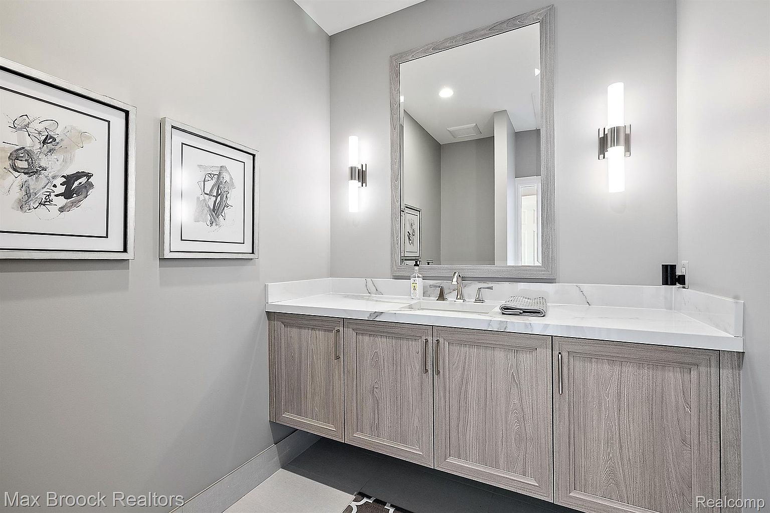 This is a well-lit guest bathroom featuring a modern vanity with a light wood finish and a white marble countertop. A large framed mirror hangs above the sink, flanked by contemporary sconces. Two framed abstract art pieces adorn the wall, adding a touch of sophistication to the space.