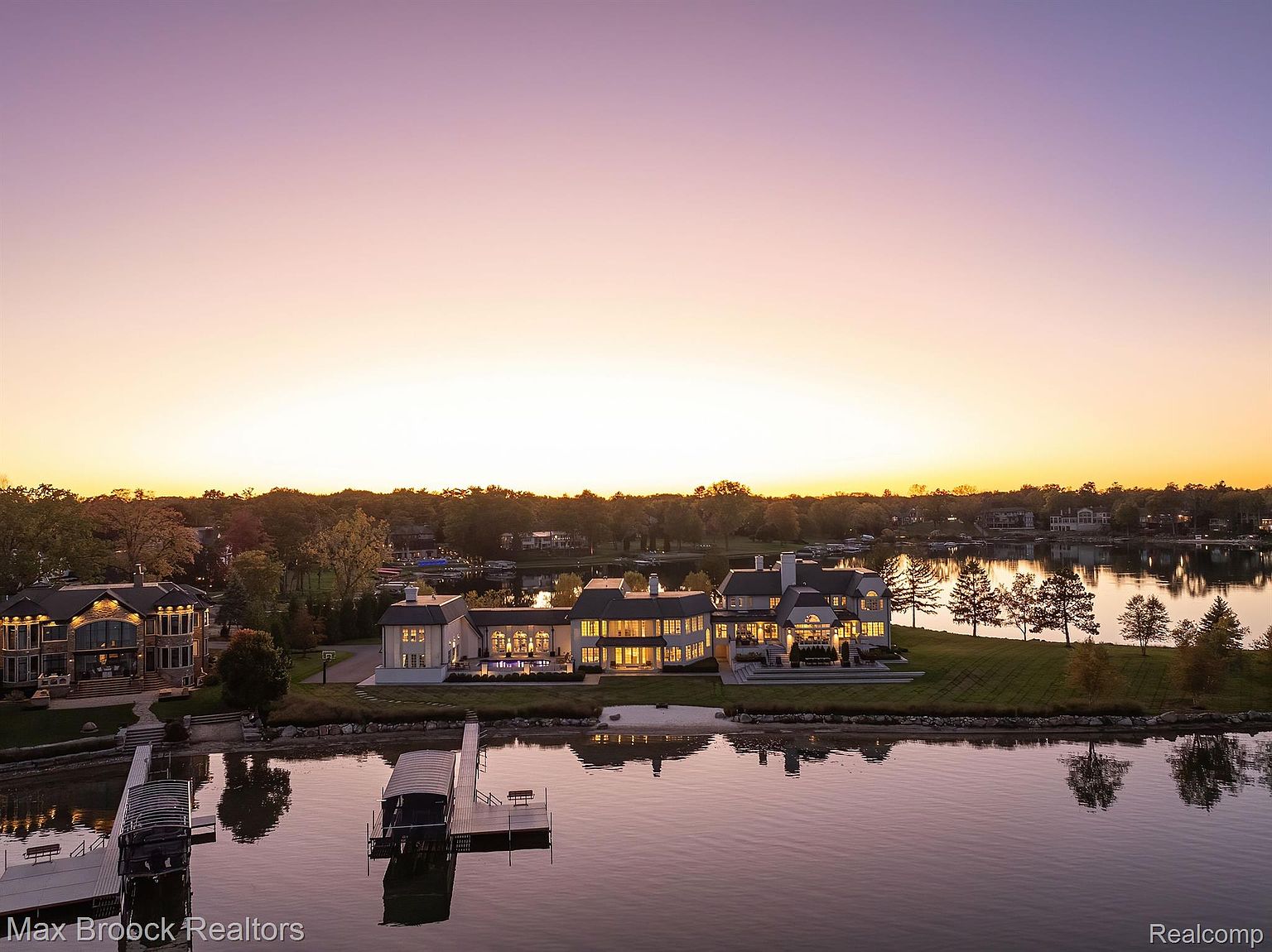 This aerial view showcases a luxurious waterfront estate at sunset. The property features multiple interconnected buildings with illuminated windows, a well-manicured lawn, and private docks extending into the calm water. The overall impression is one of opulence and serene lakeside living.