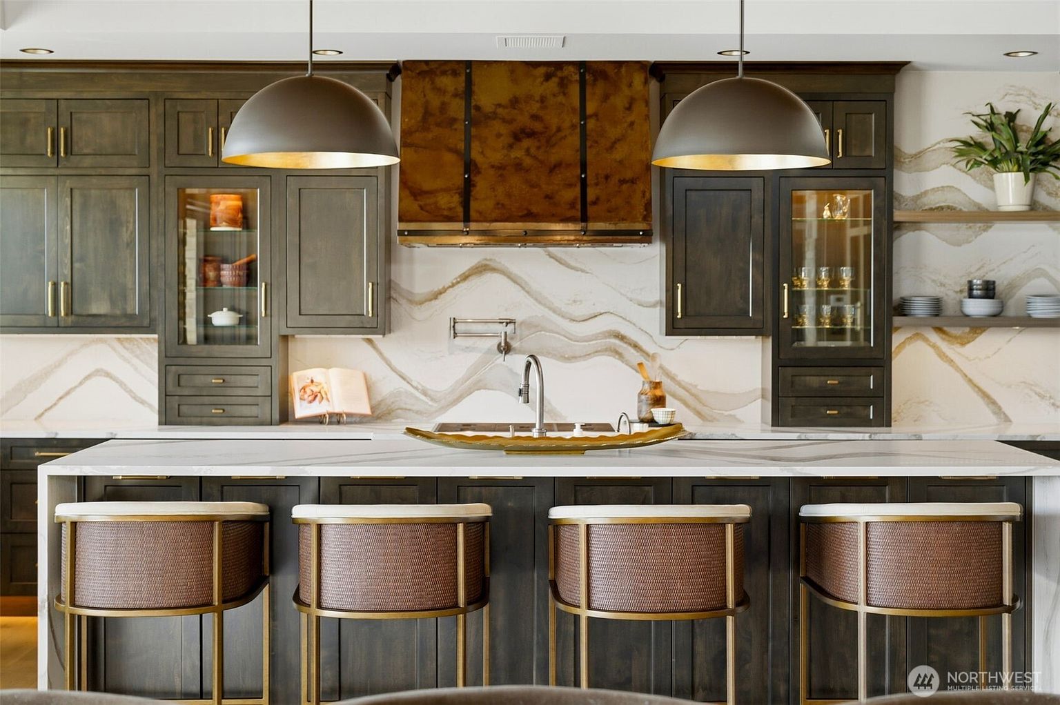 This sophisticated kitchen features dark wood cabinetry paired with a striking, wavy-patterned stone backsplash and a large central island with seating. A prominent, textured metal range hood serves as a focal point above the cooktop, complemented by two large, dome-shaped pendant lights. The space is accented with warm metallic hardware and open shelving, creating a luxurious and modern aesthetic.