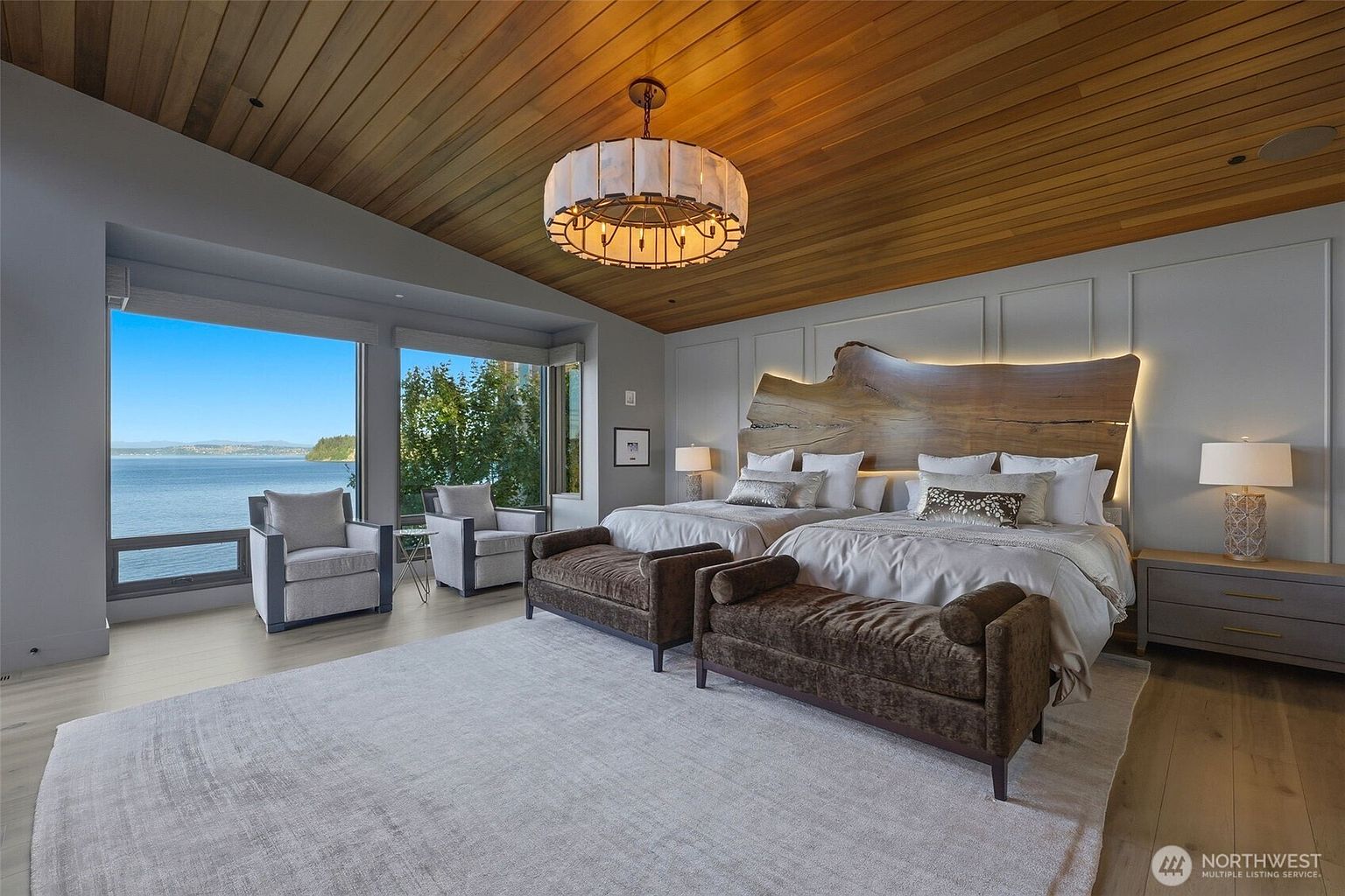 This spacious primary bedroom features a striking live-edge wood headboard with integrated backlighting, set against a wall with decorative molding. The room is anchored by two beds with matching brown upholstered benches at their feet, all resting on a large, neutral area rug. Large windows provide an expansive, unobstructed view of the water, while a warm wood-paneled ceiling and a modern chandelier add a sophisticated, natural aesthetic to the space.