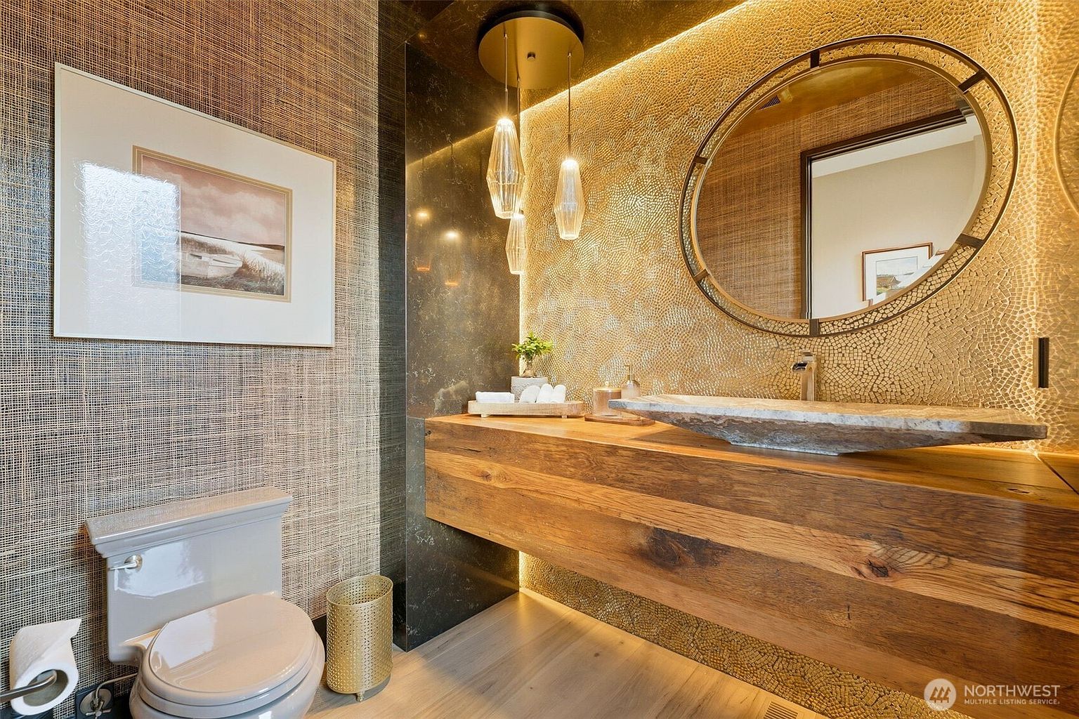 This sophisticated powder room features a floating vanity crafted from a thick slab of natural wood, topped with a rustic stone vessel sink. The walls are adorned with textured, woven-look wallpaper and a striking gold-toned mosaic accent wall that is illuminated by integrated lighting. A circular mirror, modern pendant lights, and a sleek toilet complete this high-end, contemporary space.