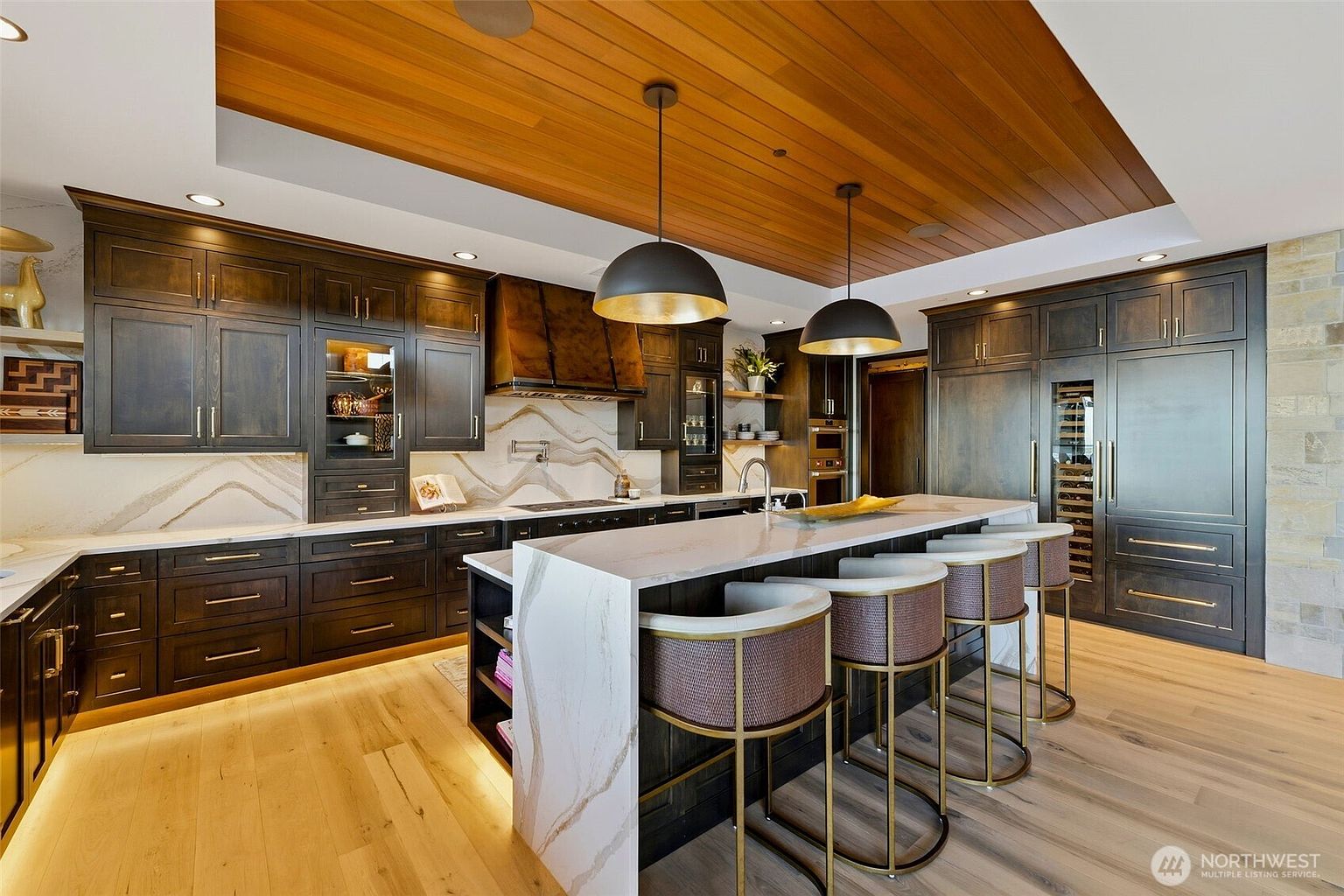 This sophisticated kitchen features dark, rich-toned cabinetry paired with a waterfall-edge island topped with light, veined stone. The space is defined by a warm wood-paneled ceiling and two large, dark pendant lights that anchor the island seating area. A stone-clad backsplash complements the overall aesthetic, while integrated appliances and a built-in wine cooler provide a seamless, high-end look.