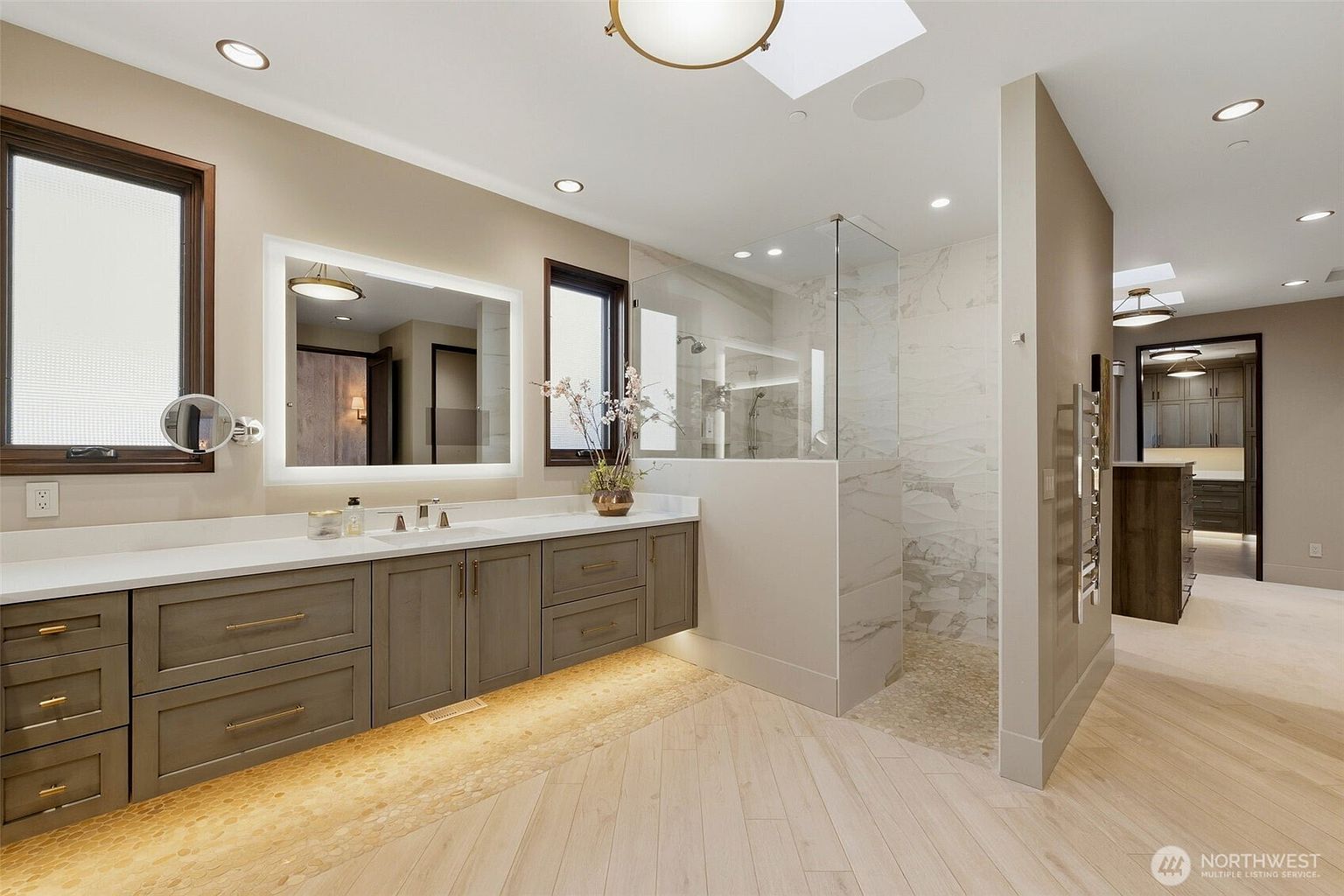 This modern primary bathroom features a floating vanity with light-toned cabinetry and a sleek, illuminated mirror. To the right, a glass-enclosed walk-in shower showcases light-colored stone tile work and a pebble-textured floor. The room is finished with light wood-look flooring laid in a herringbone pattern, creating a bright and sophisticated atmosphere.