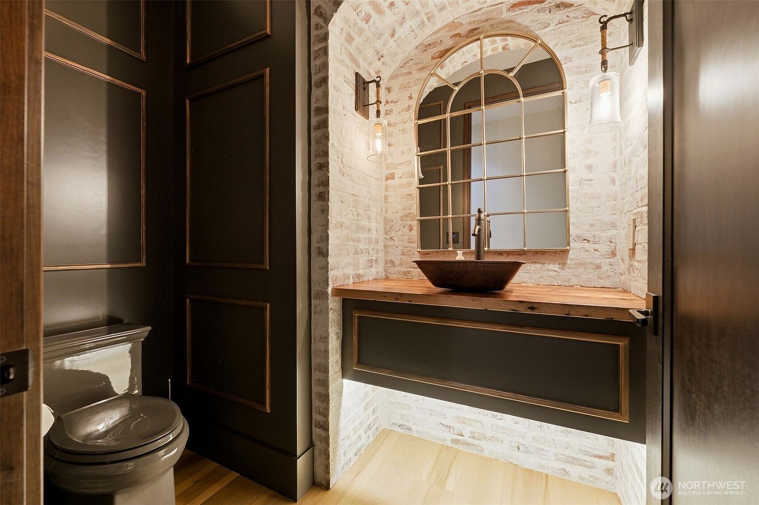 This sophisticated powder room features a striking contrast between dark, paneled walls and a rustic, white-washed brick alcove. A vessel sink sits atop a natural wood vanity, complemented by a gold-framed arched mirror and warm under-cabinet lighting. The space is finished with a dark-toned toilet and light wood flooring, creating a moody yet elegant atmosphere.