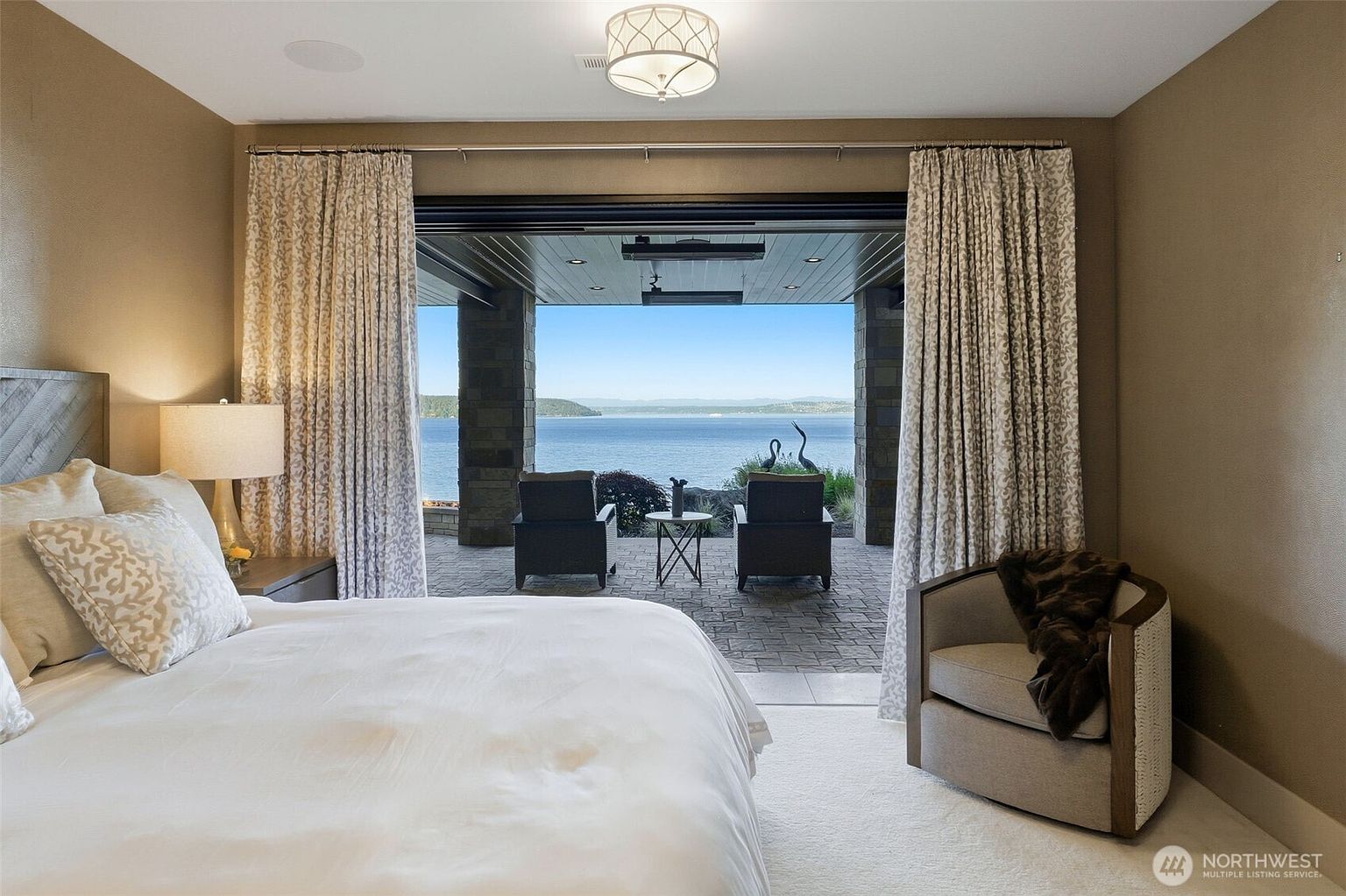 This primary bedroom features a neutral color palette and a plush white carpet, centered around a bed with patterned pillows. The room opens directly onto a covered patio through wide, retractable glass doors, providing an expansive, unobstructed view of the water and distant hills. A comfortable armchair with a throw blanket sits in the corner, creating a serene and luxurious atmosphere.