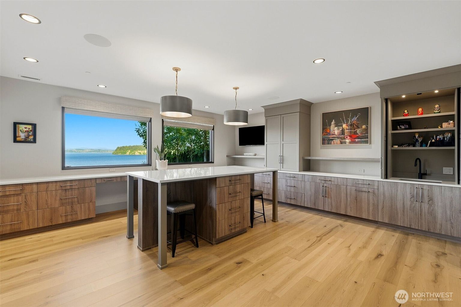 This modern kitchen features a large central island with bar seating and light-toned wood cabinetry that extends along the walls. Large windows provide a panoramic view of the water, while recessed lighting and two pendant lights illuminate the space. The room includes a built-in beverage station with a sink and open shelving, creating a clean and functional aesthetic.