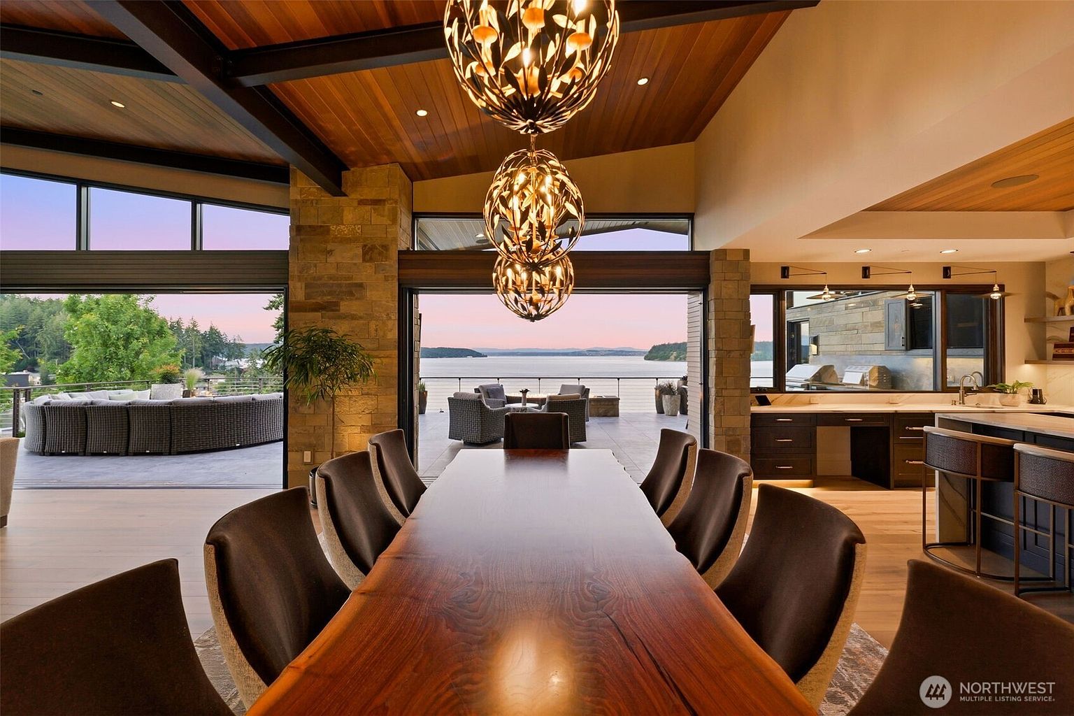 This elegant dining area features a long, natural-edge wood table surrounded by dark upholstered chairs, positioned beneath a pair of intricate, spherical pendant lights. The room seamlessly transitions to an expansive outdoor deck through wide, retractable glass walls that frame a panoramic view of the water. The space is defined by a warm wood-paneled ceiling and stone accents, creating a sophisticated, open-concept atmosphere that emphasizes indoor-outdoor living.