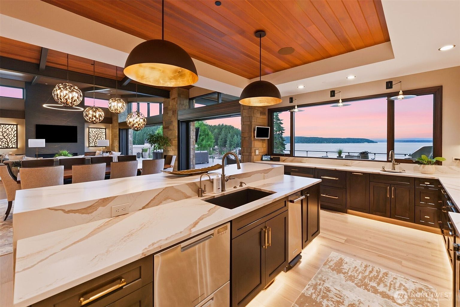 This expansive, open-concept kitchen features dark cabinetry contrasted by a large island with a light-toned stone countertop and integrated sink. The space is illuminated by prominent pendant lighting and recessed lights, while a wall of large windows provides a panoramic view of the water at sunset. The design seamlessly transitions into a dining and living area, creating a sophisticated and airy atmosphere.