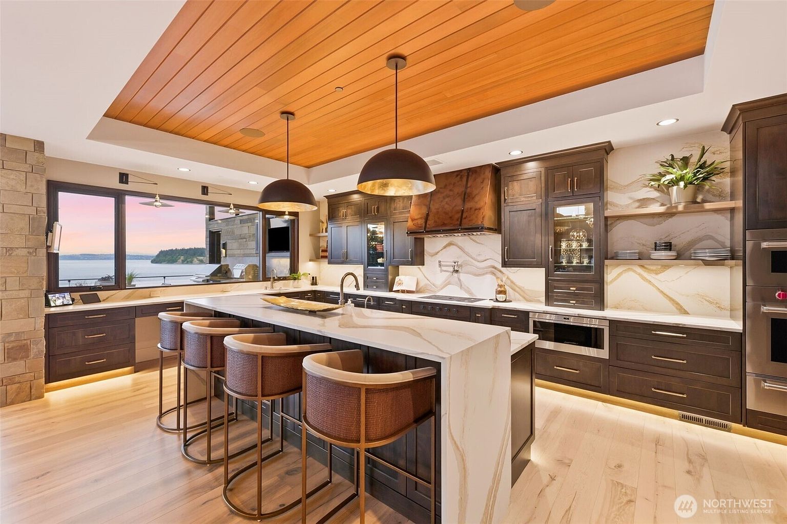 This sophisticated kitchen features a large central island with bar seating, dark cabinetry, and a striking stone backsplash with dramatic veining. The space is defined by a warm wood-paneled ceiling, pendant lighting, and expansive windows that provide a scenic view of the water. The overall aesthetic is modern and luxurious, blending rich textures with a clean, open layout.