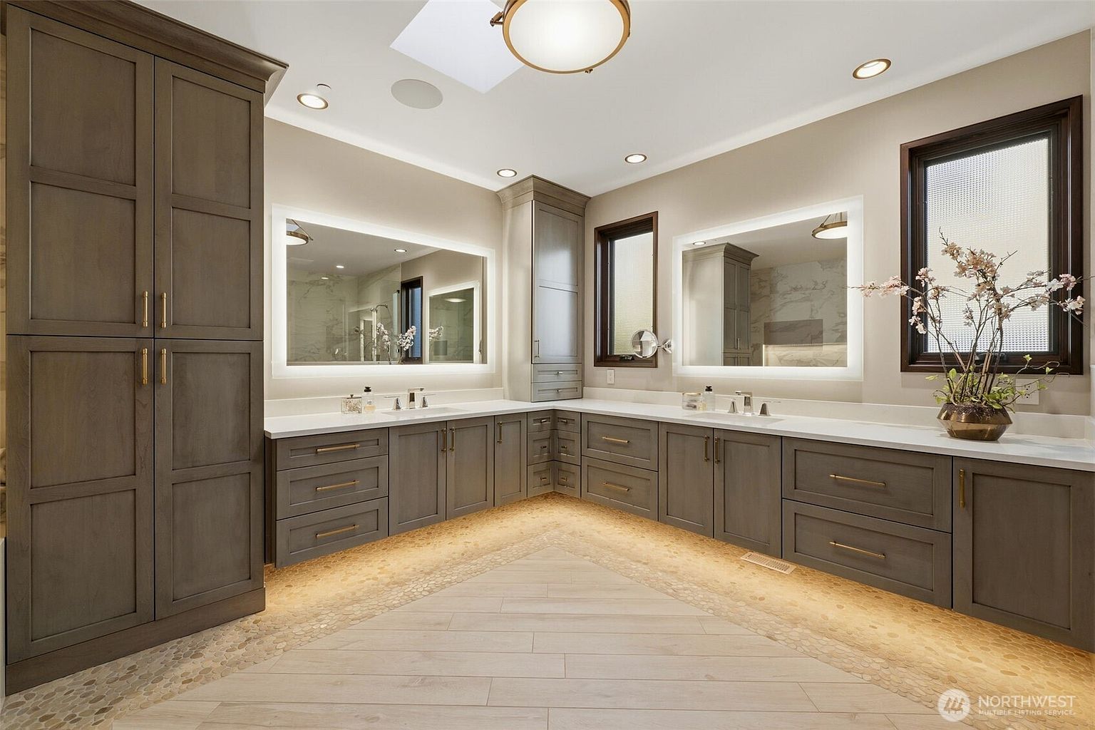 This spacious primary bathroom features a large L-shaped vanity with dark-toned cabinetry, gold-toned hardware, and a light-colored countertop. Two illuminated mirrors are mounted above the sinks, complemented by recessed lighting and a skylight that brightens the room. The flooring is a unique combination of light-toned wood-look planks bordered by a decorative pebble-style inlay, creating a sophisticated and modern aesthetic.