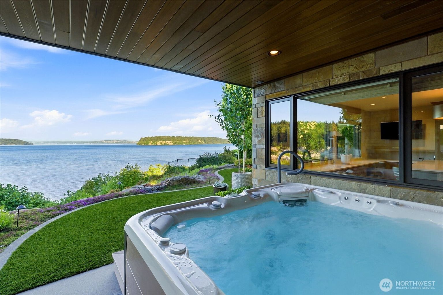 This image captures a luxurious outdoor spa area set on a covered patio, offering a panoramic view of a serene bay and distant forested hills. The spa features built-in seating and is positioned adjacent to a stone-clad wall and large windows that provide a glimpse into the home's interior. A manicured lawn and lush landscaping border the patio, creating a seamless transition between the private relaxation space and the natural waterfront surroundings.