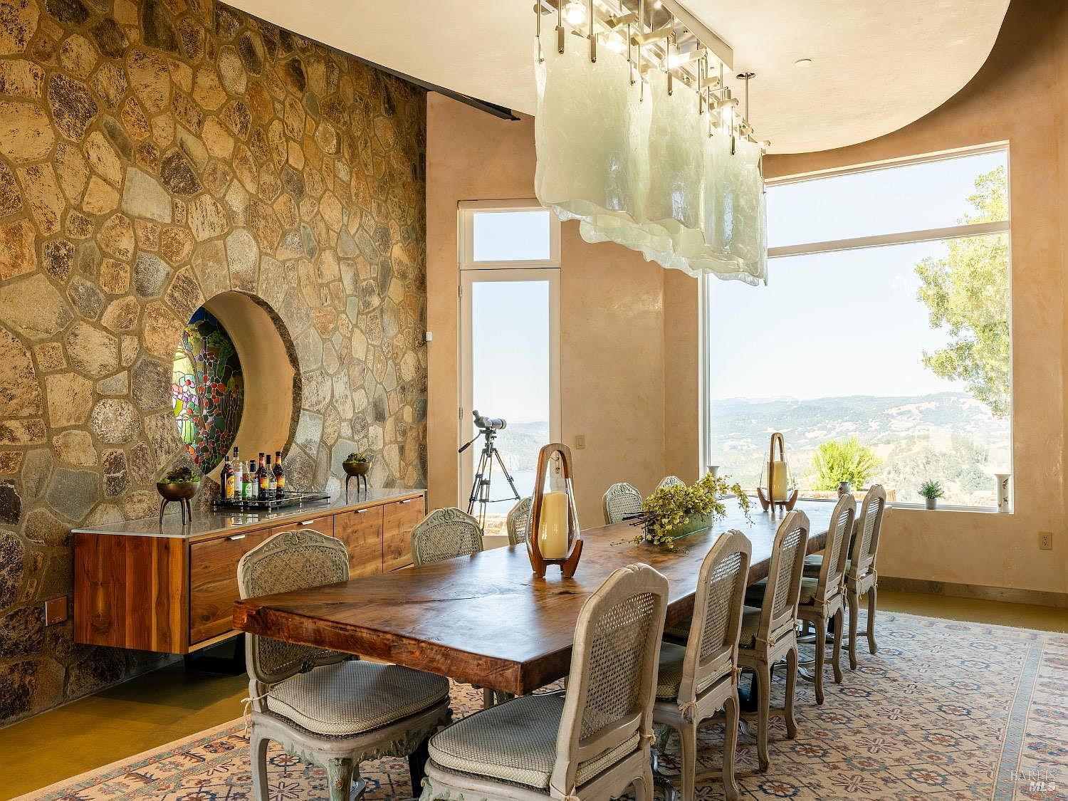 This elegant dining room features a large, rustic wooden table surrounded by upholstered chairs, set against a striking stone-clad wall with a circular stained-glass accent. A unique, textured glass chandelier hangs above the table, while expansive windows provide a panoramic view of the surrounding landscape. The room is anchored by a patterned area rug and a long, low-profile wooden sideboard, creating a sophisticated and inviting atmosphere.