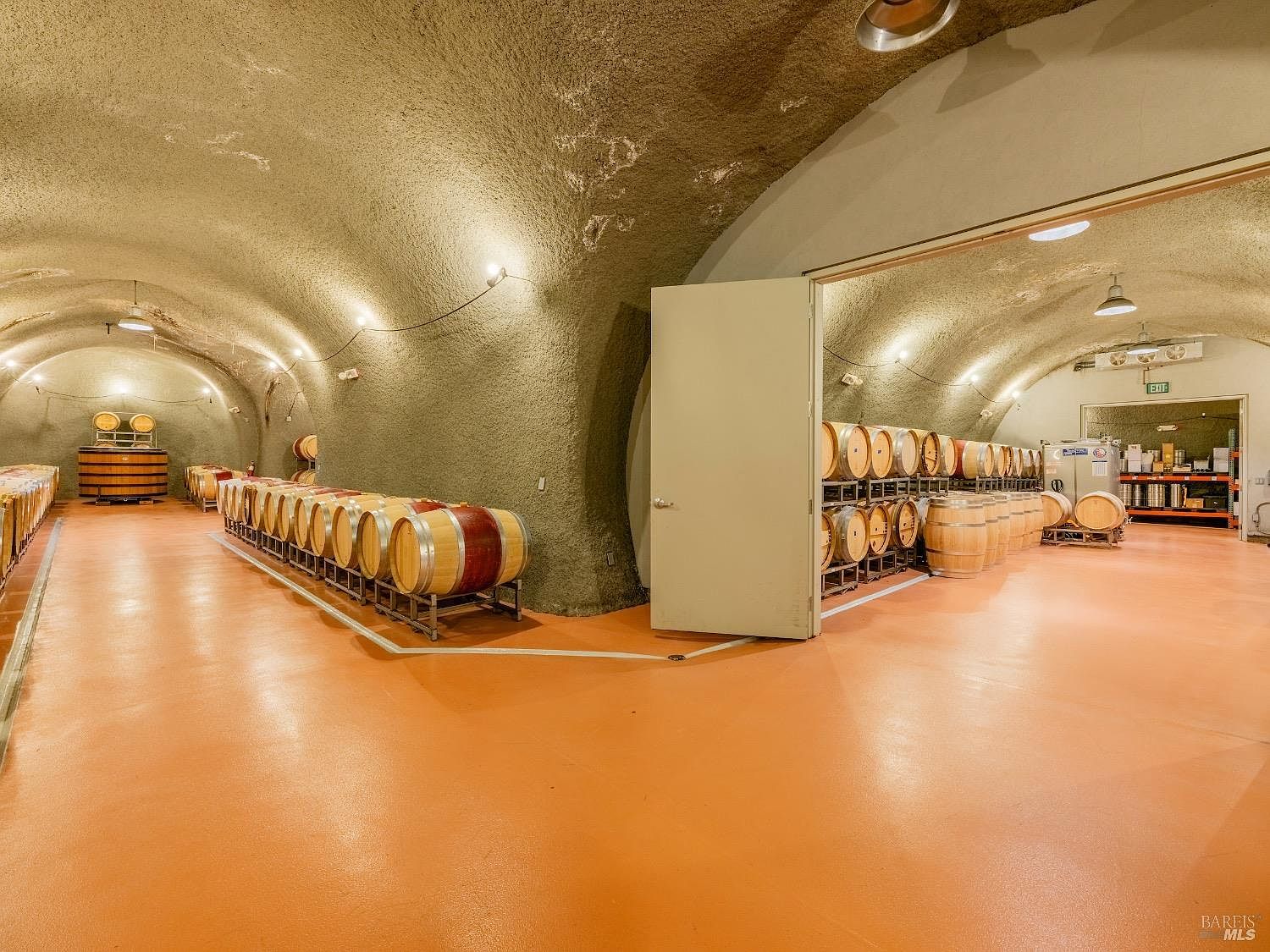 This expansive wine cellar features a vaulted, textured ceiling and a smooth, orange-toned floor, creating a unique and industrial aesthetic. Rows of wooden wine barrels are neatly arranged on metal racks throughout the space, which is divided into two distinct sections by an open doorway. The perspective captures the depth of the cellar, emphasizing the organized storage capacity and the cavernous, climate-controlled atmosphere.