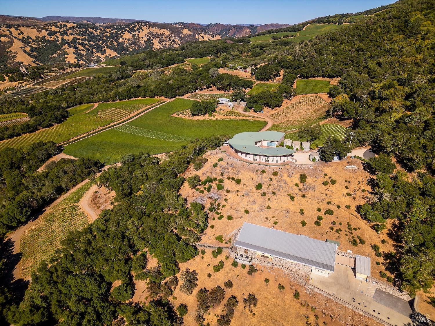 This high-angle aerial view captures a sprawling rural estate nestled within a hilly, vineyard-covered landscape. The property features a distinctive curved residence perched on a ridge, complemented by a large, rectangular stone-clad outbuilding situated lower on the slope. The surrounding terrain is a mix of cultivated grapevines, native oak trees, and dry, golden-hued hills, emphasizing the expansive and private nature of the estate.