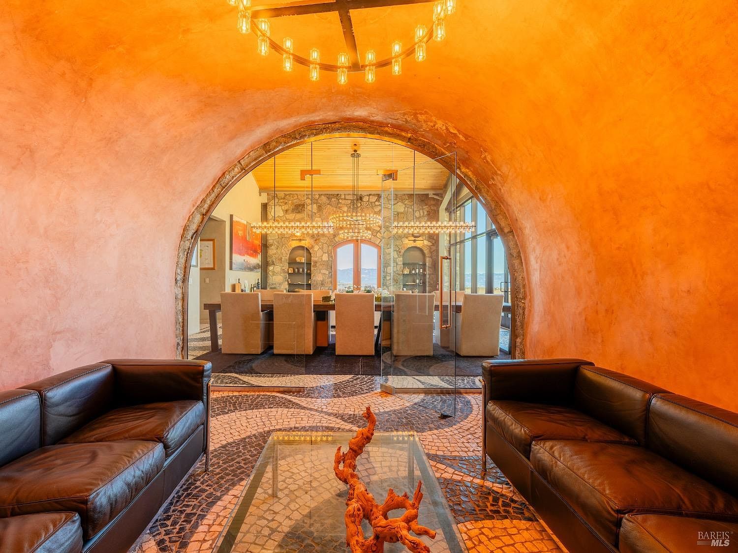 This interior view captures a sophisticated living area that transitions into a formal dining room through a dramatic, arched stone entryway. The foreground features two dark leather sofas flanking a glass-topped coffee table with a decorative driftwood centerpiece, all set upon a striking mosaic-tiled floor. Beyond the glass partition, the dining space is anchored by a long table and stone-clad walls, creating a warm, textured atmosphere illuminated by elegant circular chandeliers.