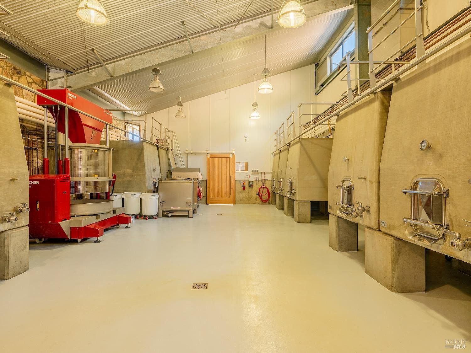 This image depicts a professional-grade winery production facility featuring large industrial fermentation tanks and a prominent red Bucher Vaslin grape press. The space is characterized by high, vaulted ceilings with exposed metal beams, industrial lighting fixtures, and a clean, light-colored concrete floor. The perspective is a wide-angle shot looking down the center aisle, showcasing the functional and utilitarian design of a commercial winemaking operation.
