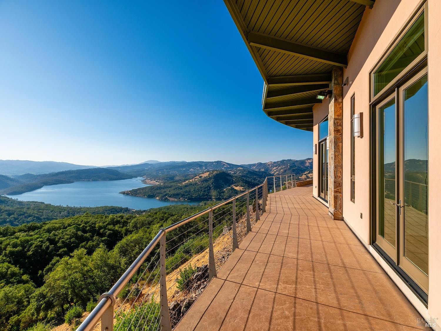 This expansive, elevated deck offers a panoramic view of a large lake surrounded by rolling, tree-covered hills under a clear blue sky. The deck features a smooth, neutral-toned surface and a modern metal railing with thin cables, providing an unobstructed sightline to the landscape. Large glass doors open from the home onto the deck, which is sheltered by a wide, angled roof overhang with recessed lighting.