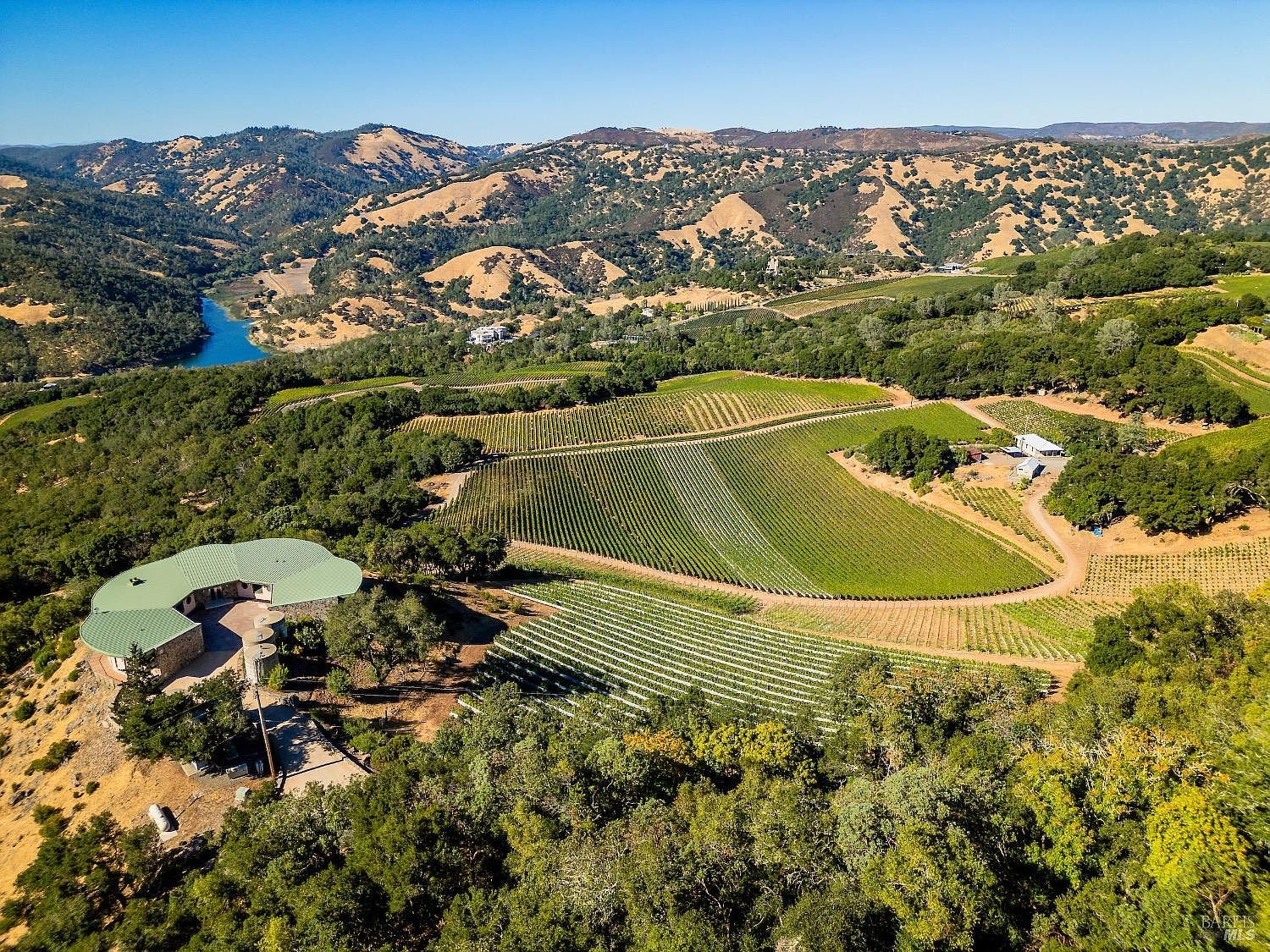 This expansive aerial view captures a sprawling estate nestled within a rolling, sun-drenched landscape of vineyards and native oak trees. A unique, curved residence with a green roof sits prominently on a hillside, overlooking the meticulously maintained rows of grapevines and a distant blue reservoir. The scene conveys a sense of serene, rural luxury, highlighting the property's vast acreage and integration with the natural topography.