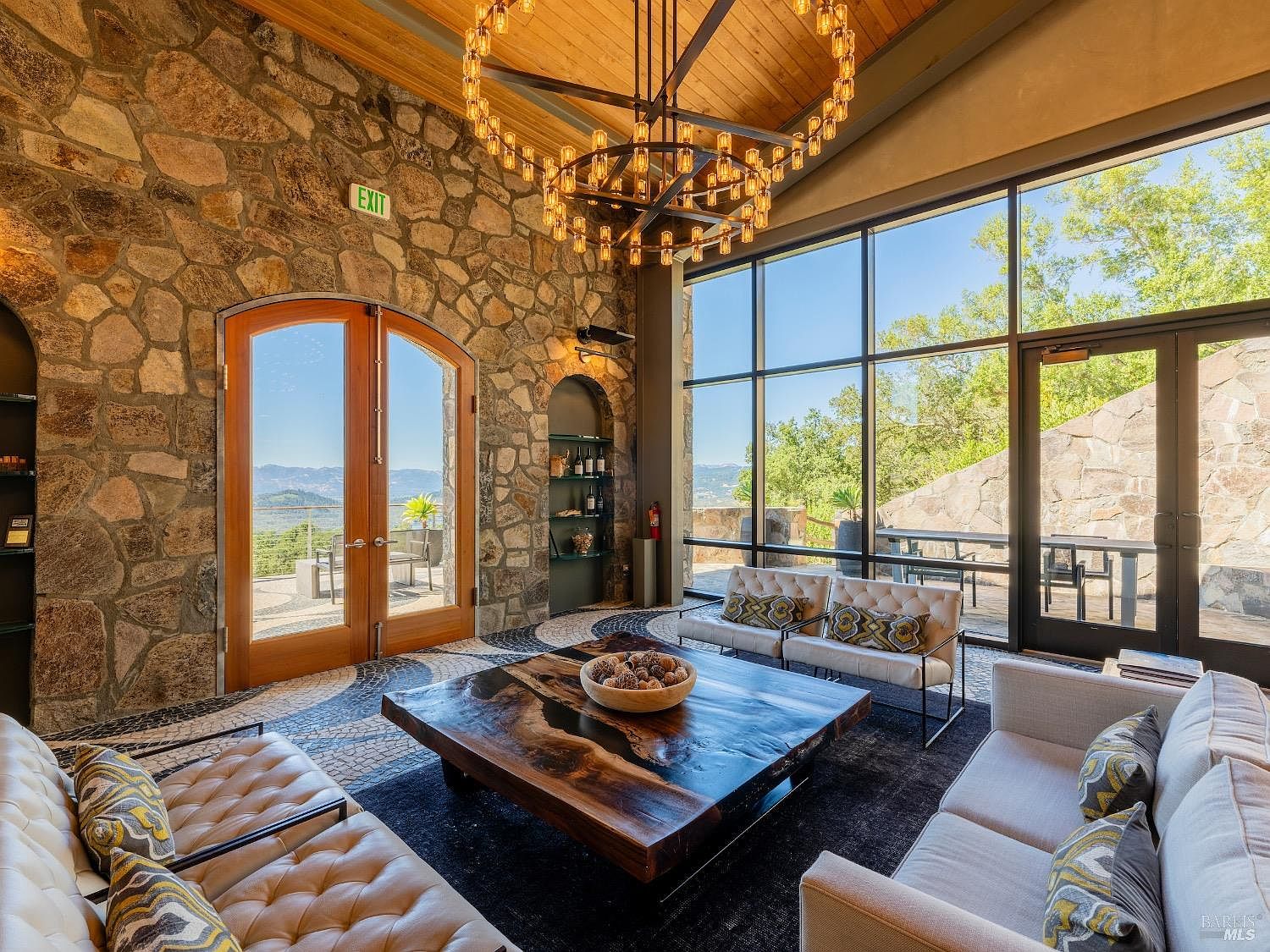 This sophisticated living room features a dramatic stone wall, a large circular chandelier, and a unique live-edge wood coffee table as the centerpiece. The space is framed by expansive glass walls that offer panoramic views of the surrounding hills and provide abundant natural light. The combination of rustic stone textures, warm wood ceilings, and modern furniture creates an elegant and inviting atmosphere.