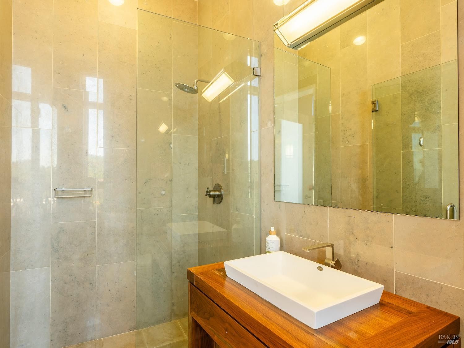 This modern bathroom features a floating wood vanity topped with a rectangular vessel sink and a sleek, wall-mounted faucet. To the left, a glass-enclosed walk-in shower is finished with neutral-toned stone tiles, while a large, frameless mirror hangs above the vanity to enhance the sense of space. The room is illuminated by a minimalist light fixture, creating a clean and contemporary aesthetic.
