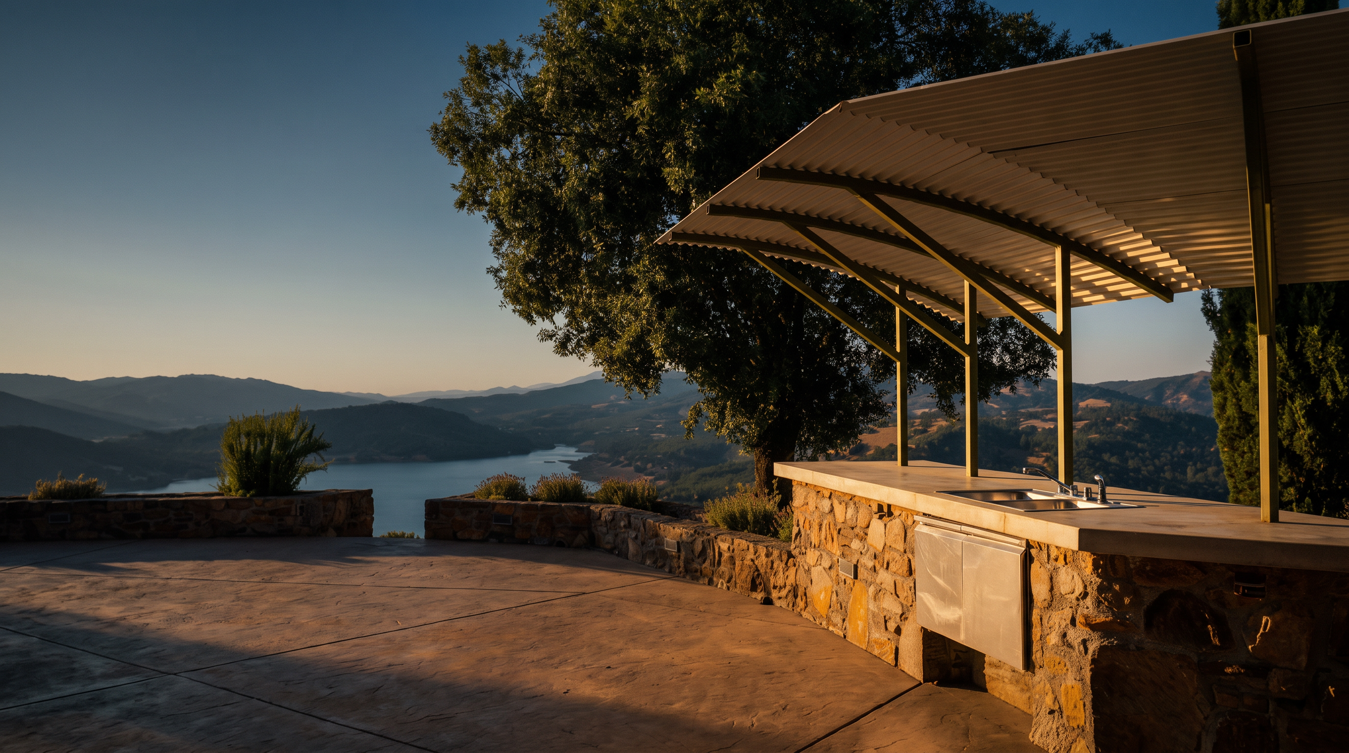 This expansive outdoor patio features a custom stone-built kitchen area sheltered by a modern, angled metal roof structure. The space offers a commanding, elevated view of a serene lake surrounded by rolling hills and lush landscape. The combination of rustic stone masonry and the open-air design creates a sophisticated yet relaxed setting perfect for outdoor entertaining.