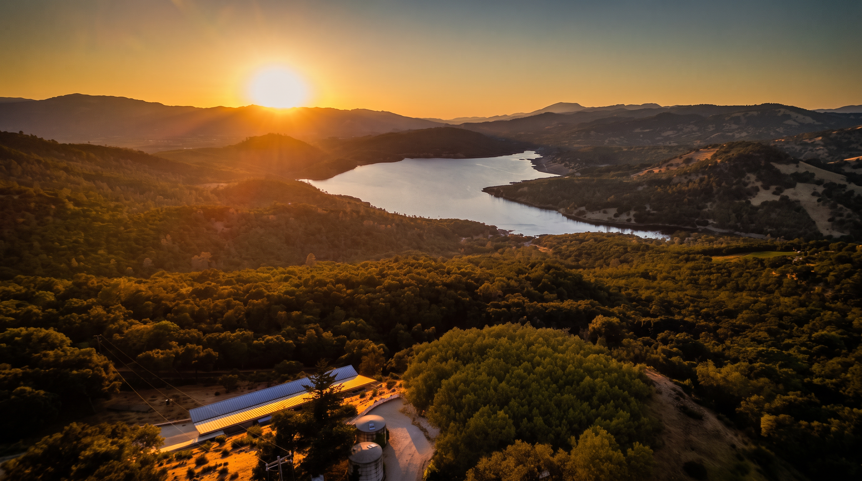 This stunning aerial perspective captures a unique, crescent-shaped residence nestled on a hillside overlooking a serene lake at sunset. The home features a distinctive metal roof and stone exterior walls, complemented by a central courtyard containing several cylindrical water storage tanks. The surrounding landscape is lush with dense forest, creating a private and tranquil setting that emphasizes the property's connection to nature.