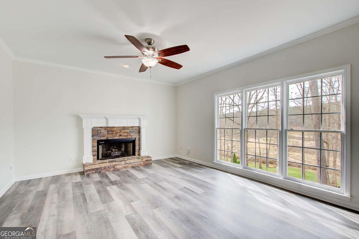 This spacious living room features light-toned wood-look flooring and neutral walls, creating a bright and airy atmosphere. A prominent stone fireplace with a white mantel serves as the focal point, while a large bank of windows provides ample natural light and a view of the outdoors. The room is finished with a ceiling fan, offering a clean and versatile space ready for furniture arrangement.