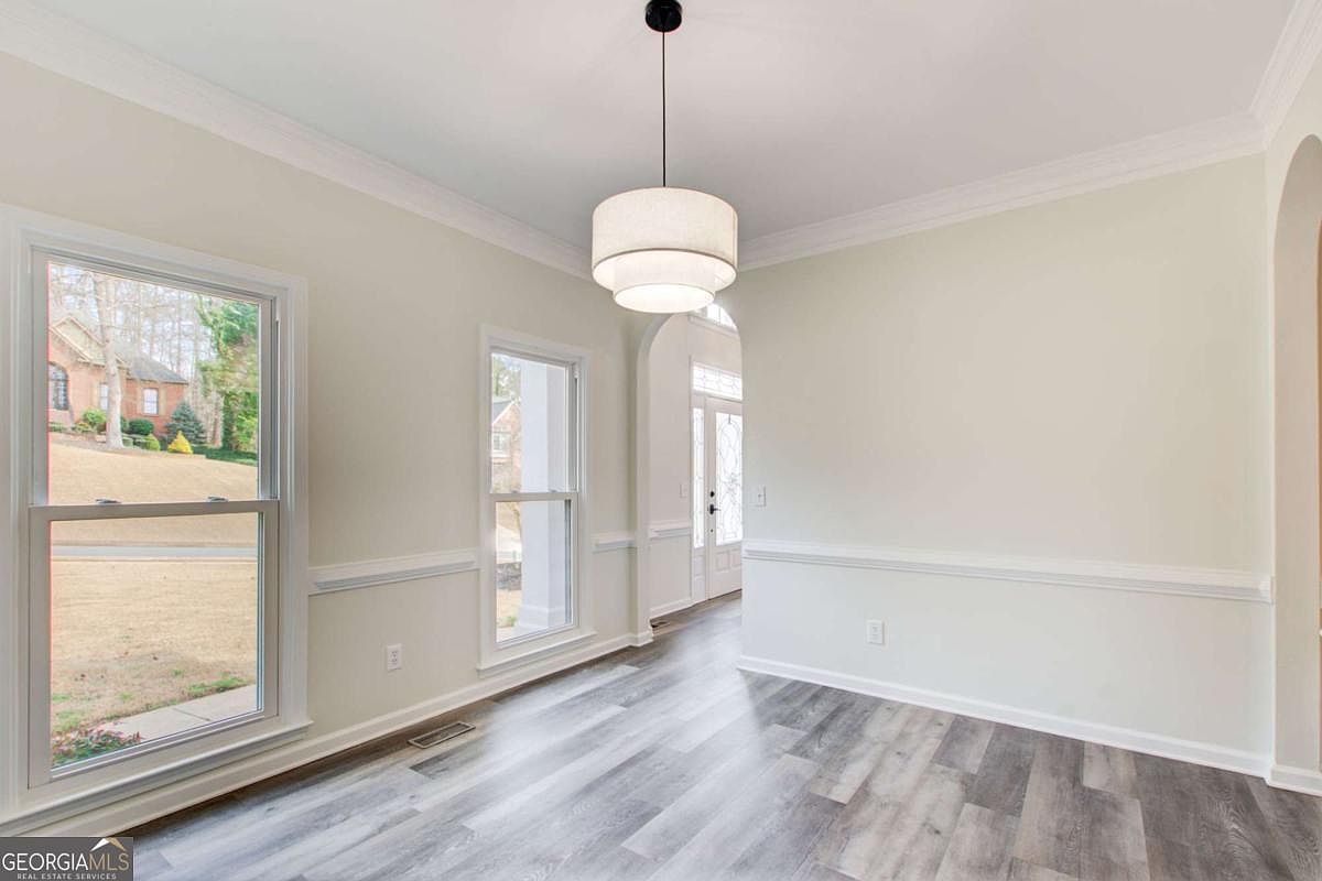 This bright and airy dining room features modern wood-look flooring and neutral wall tones, creating a clean and inviting atmosphere. A contemporary tiered pendant light fixture serves as a focal point, while large windows allow for ample natural light and views of the exterior. The space is framed by elegant crown molding and chair rail trim, offering a versatile area for formal or casual dining.