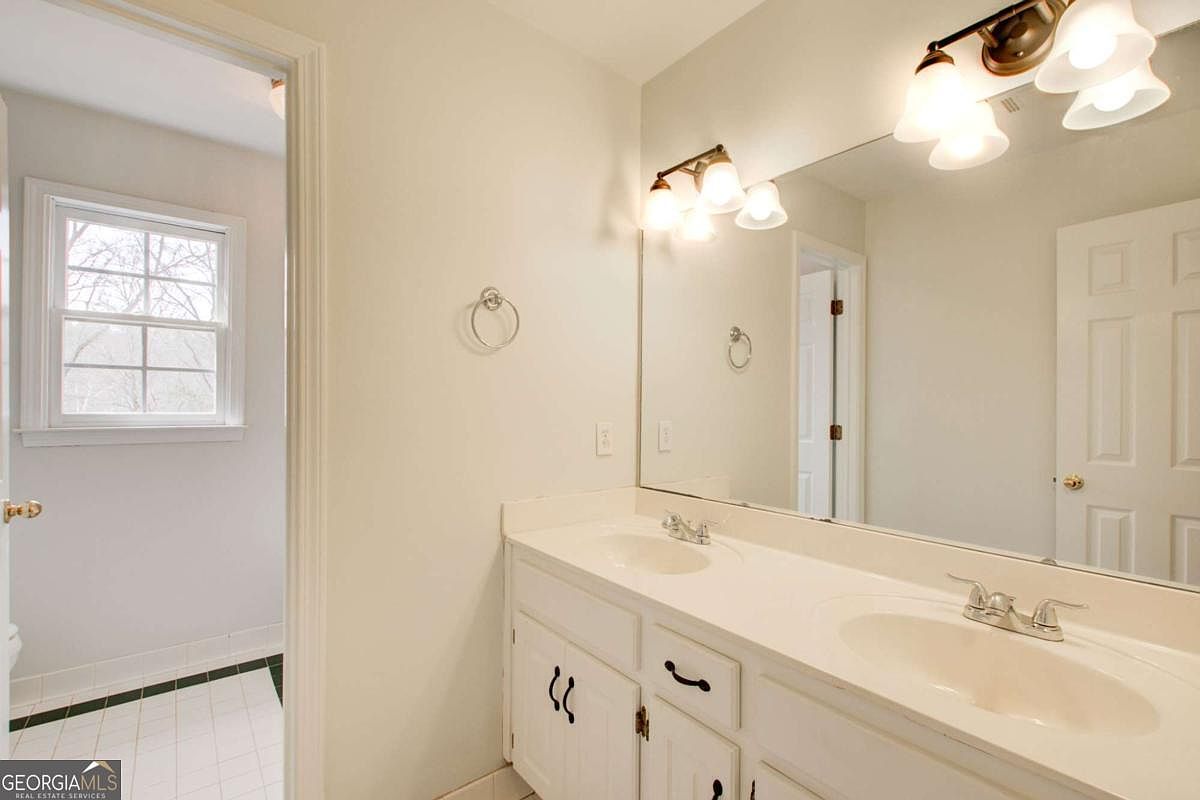 This bathroom features a double-sink vanity with white cabinetry and a large, full-width mirror that enhances the sense of space. The room is brightly lit by two multi-light fixtures and includes a doorway leading to a separate area with a window. The neutral color palette and clean lines create a functional and well-maintained appearance.