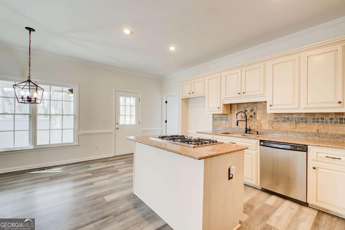 This bright and airy kitchen features cream-colored cabinetry, granite countertops, and a central island equipped with a gas cooktop. The space is finished with light-toned wood-look flooring and a decorative tile backsplash, creating a warm and inviting atmosphere. A large window and a glass-paneled door provide ample natural light, highlighting the clean, functional layout.